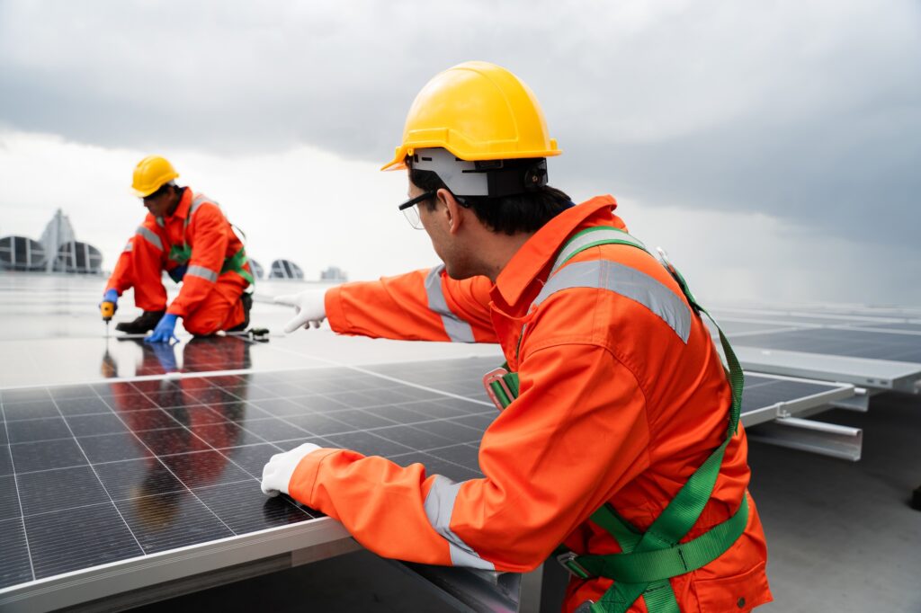 Two men in orange safety gear are working on a solar panel